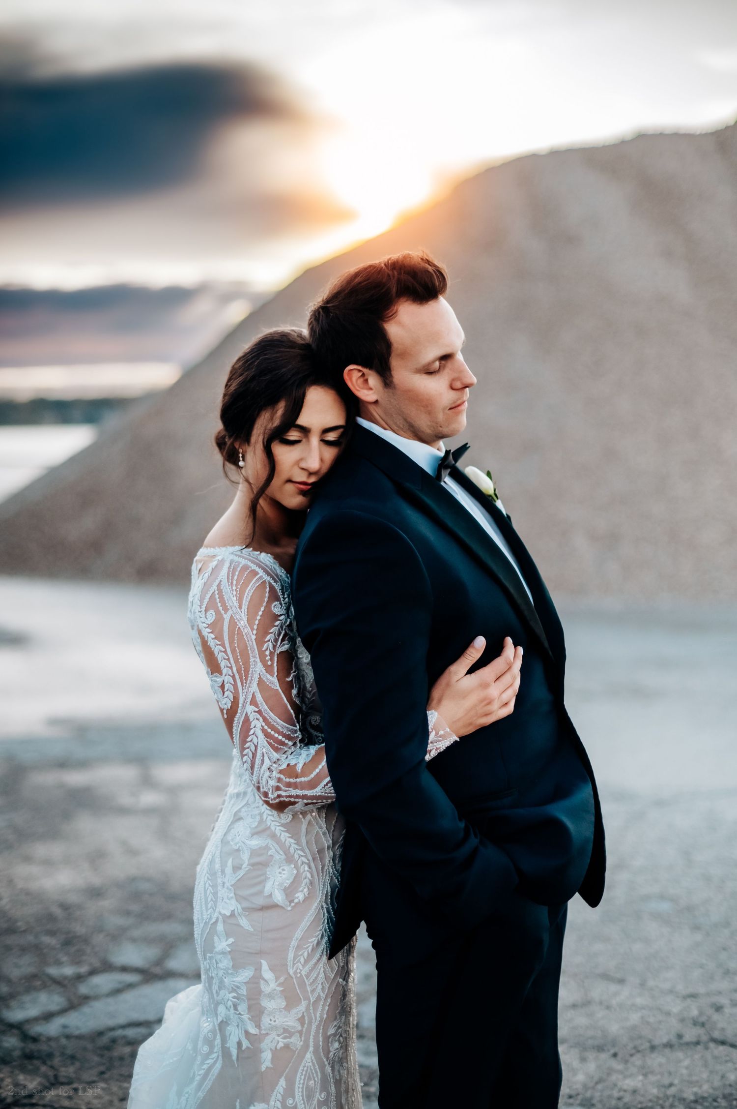 Bride and groom embrace at sunset, wearing elegant lace wedding gown and black tuxedo, with golden light behind them.
