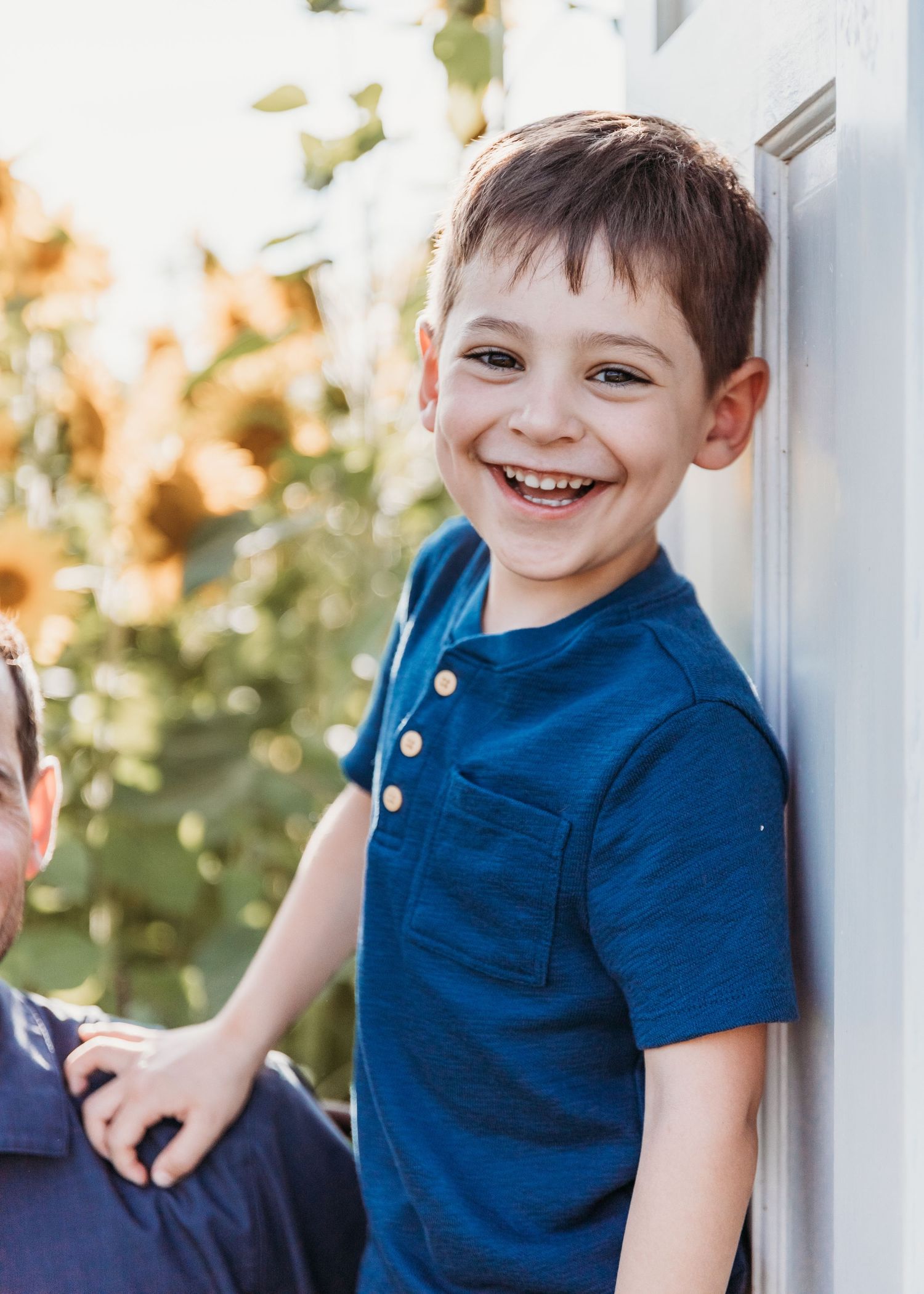 A happy young boy in a blue henley shirt laughs while leaning against a white wall outdoors.