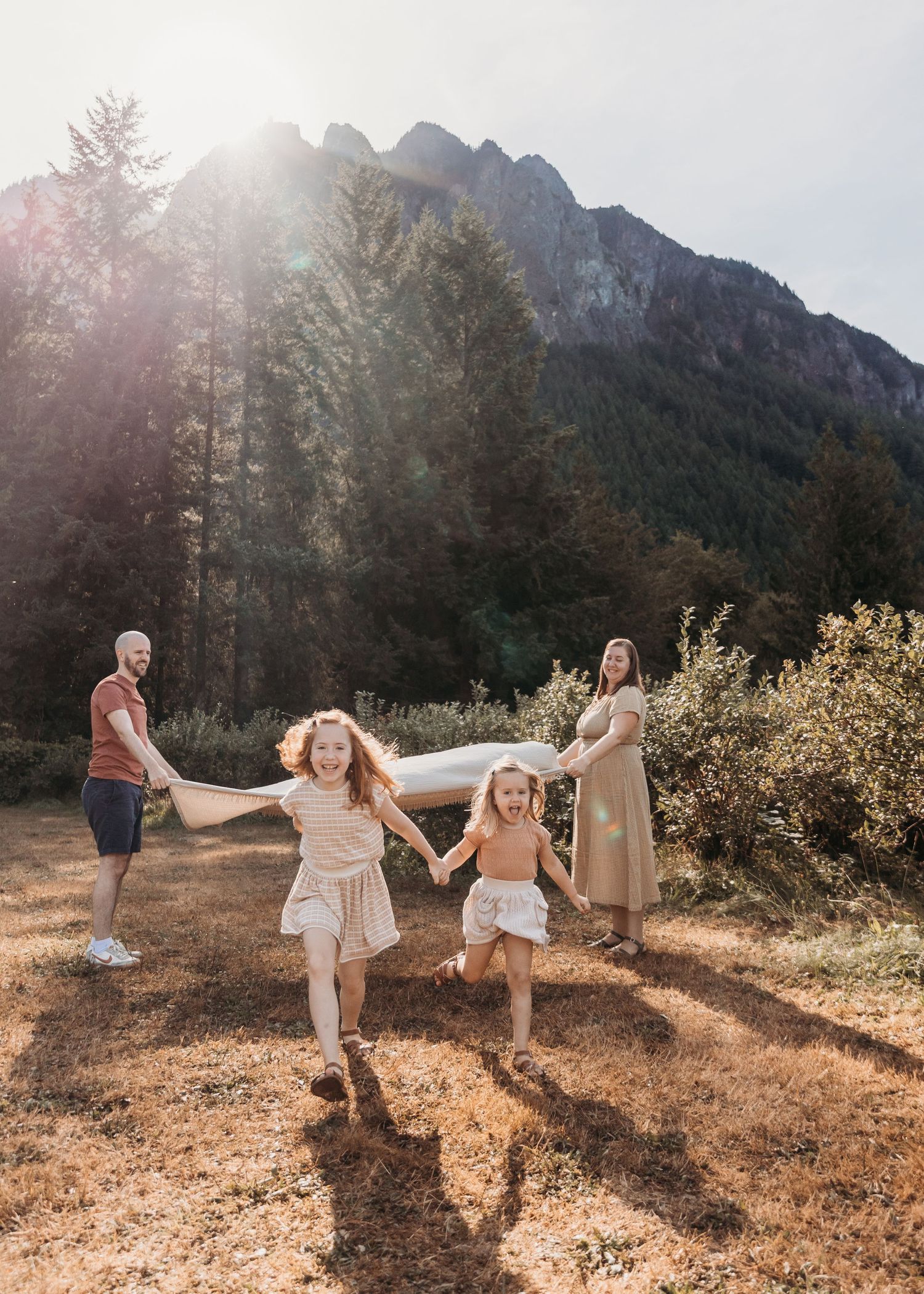 Family of four joyfully running in a sunlit mountain meadow surrounded by trees, two young girls holding hands in foreground.