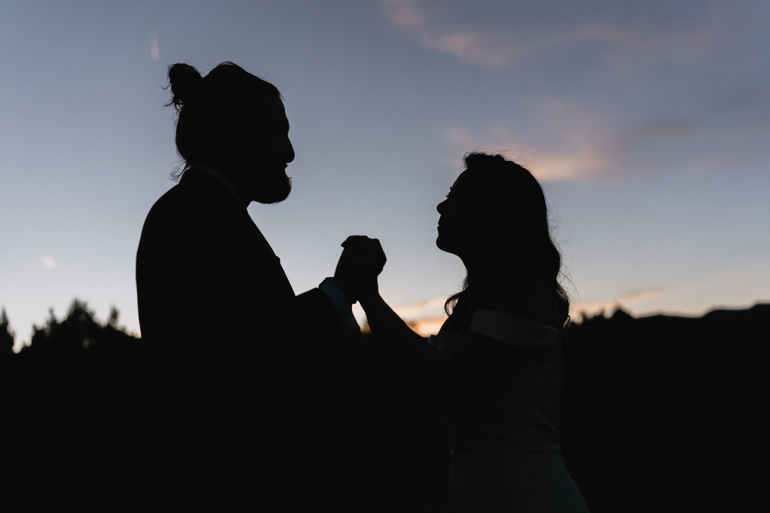 Romantic silhouette of bride and groom holding hands dancing against a twilight sky at sunset.