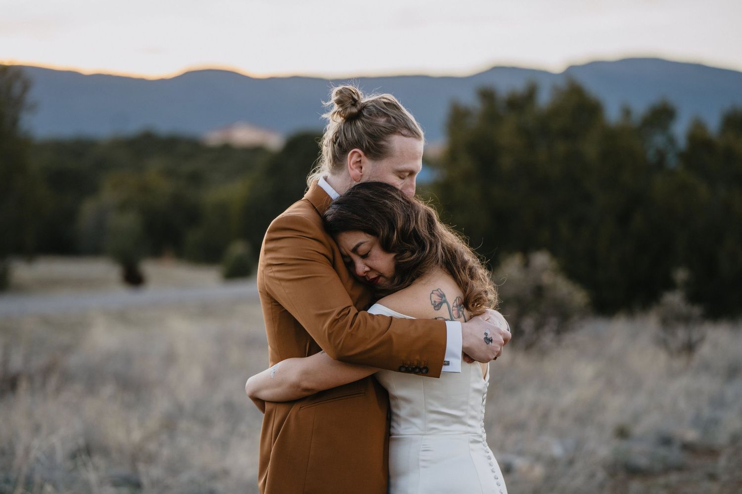 Groom in brown suit tenderly embraces bride in white dress in scenic mountain meadow at dusk.