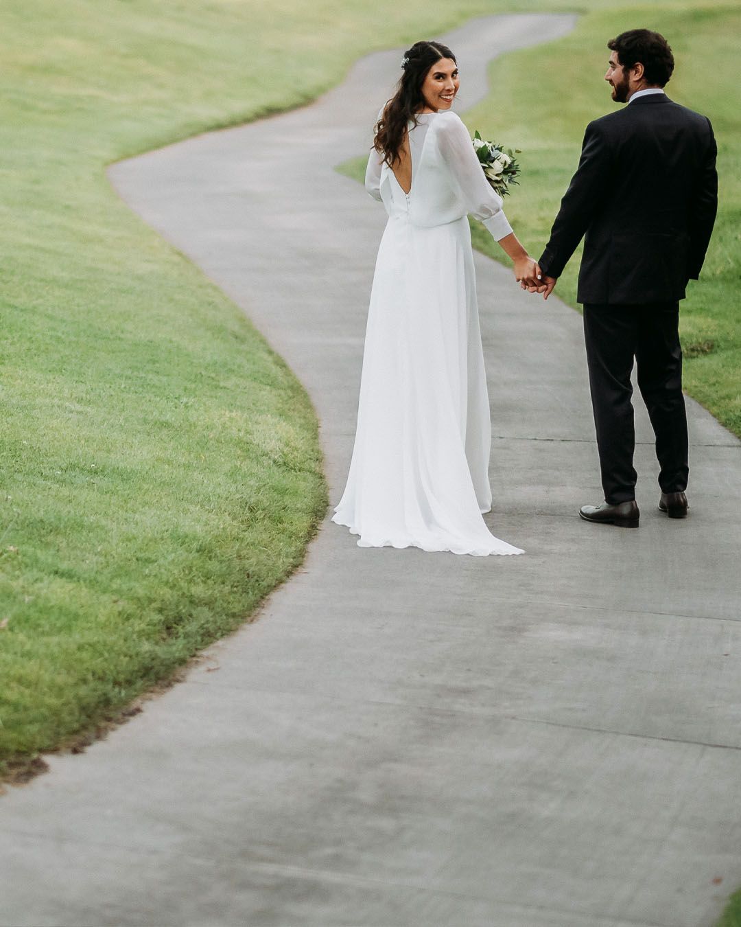 Bride and groom walking hand in hand along a winding garden path, bride glancing back.
