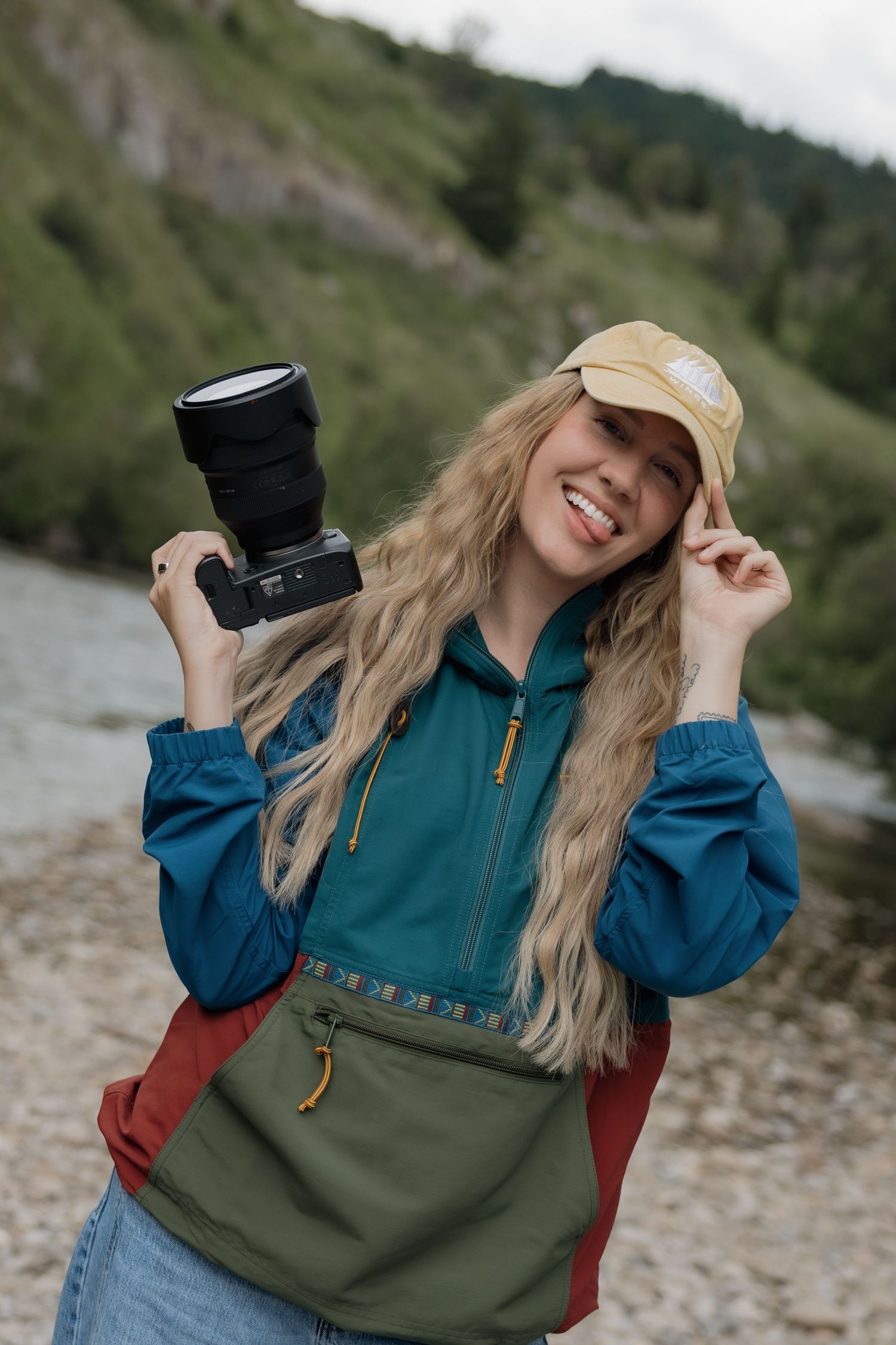 Young woman in denim shirt sits on wooden chair in shallow water, holding camera, with mountains in background.