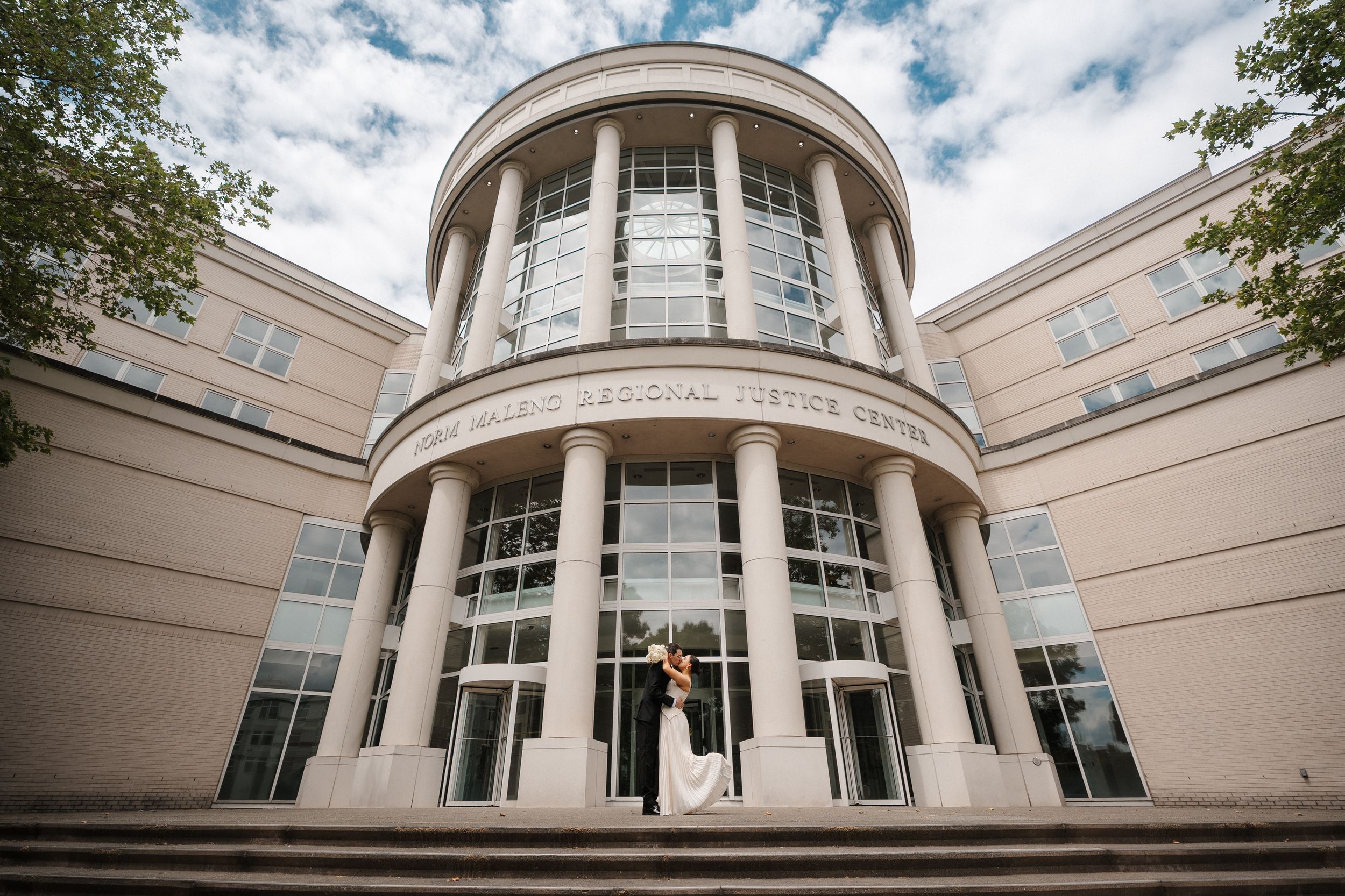 Bride and groom pose on steps of a grand beige civic building with columns and large windows.