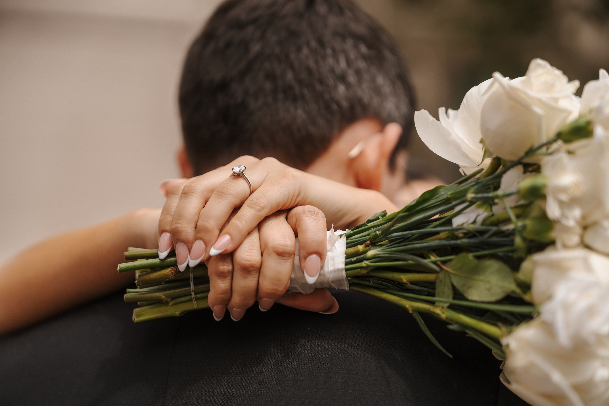 Close-up of bride and groom embracing, showing engagement ring and white rose bouquet in her hands.