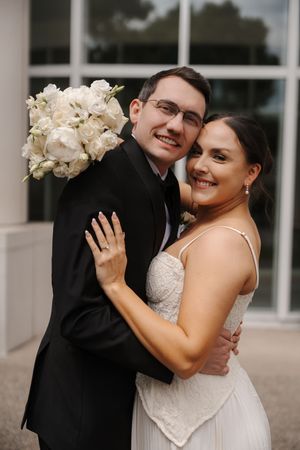 Smiling bride and groom embracing outdoors, bride holding white rose bouquet in lace gown and groom in black tuxedo.