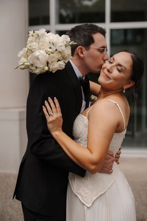 Groom in black tuxedo kissing bride on cheek as she smiles, holding white floral bouquet outside.