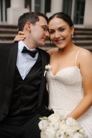 A smiling bride in a white strapless gown holds white roses as a groom in a black tuxedo nuzzles her cheek.