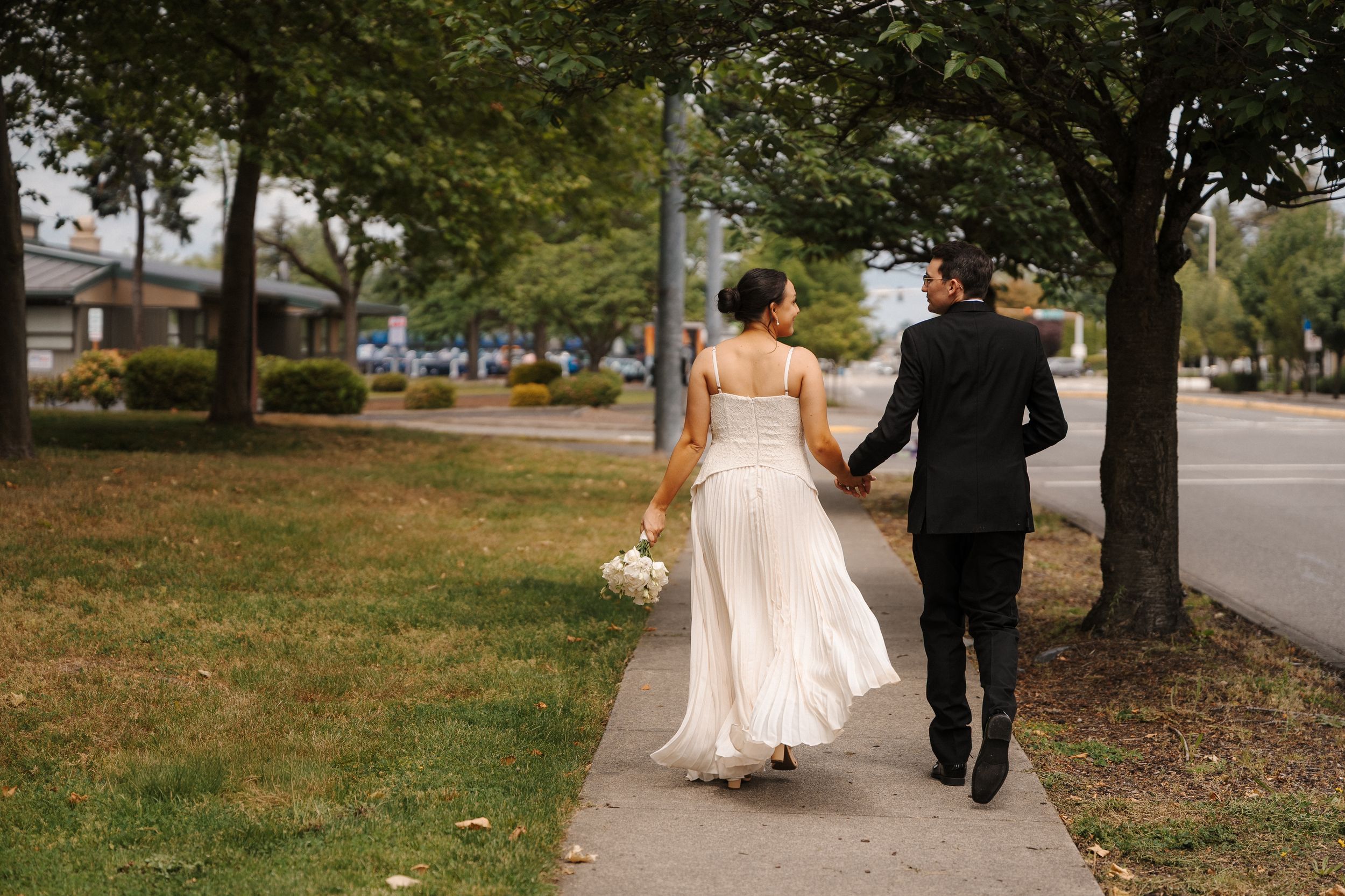 Bride and groom walking hand in hand on a tree-lined sidewalk, bride holding a white bouquet in a flowing gown.