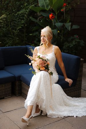 Elegant bride in white lace gown holding a pink bouquet, seated on a blue outdoor sofa surrounded by lush greenery.
