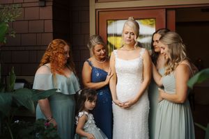 Bride in white gown surrounded by bridesmaids in sage green dresses and flower girl during wedding preparation.