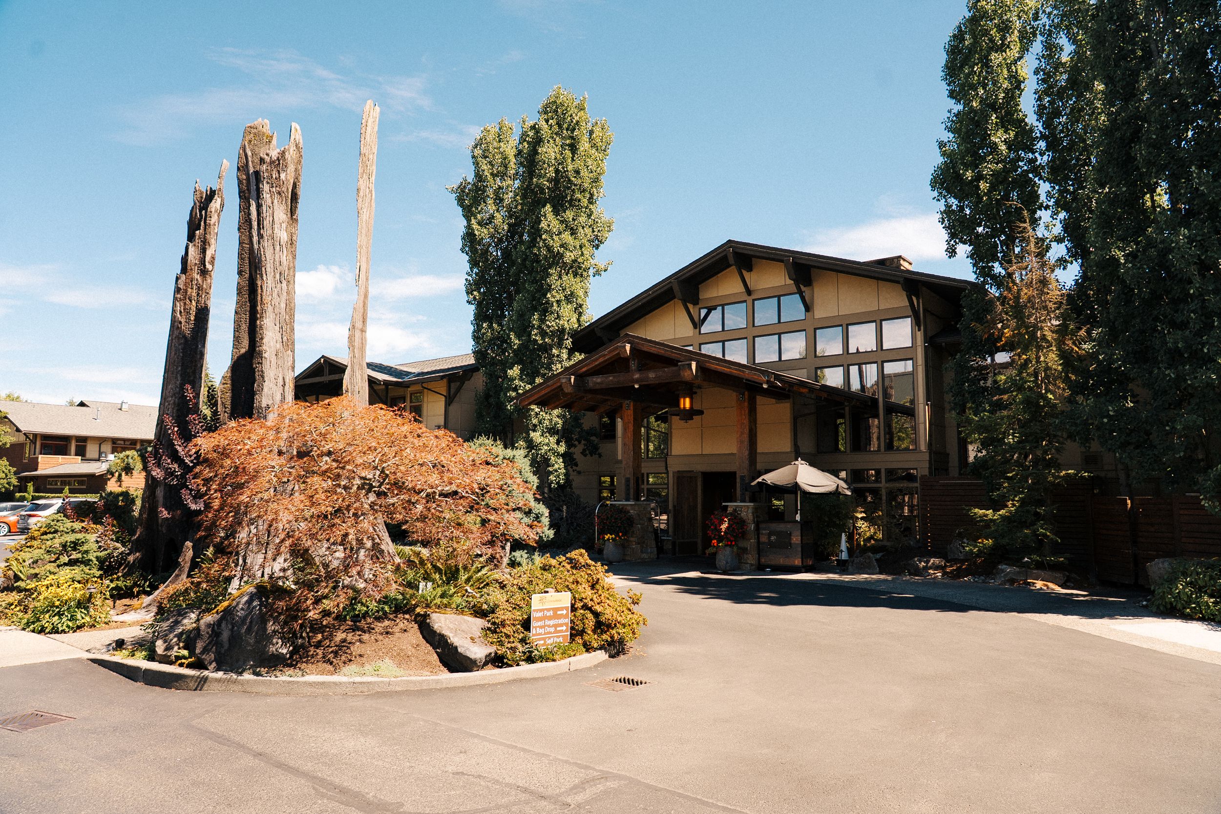 Rustic lodge-style wedding venue exterior with tall tree stumps, lush landscaping, and a paved driveway under blue skies.