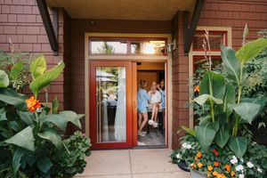 Women gathering at a red front door surrounded by lush tropical plants and colorful flowers at a home entrance.