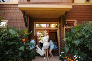 Bride in white gown entering venue doorway assisted by bridesmaids in blue, surrounded by lush tropical garden plants.