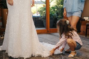 A flower girl crouches to adjust the bride's white lace wedding dress train while a woman in denim shorts stands nearby.