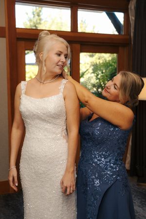 Bride in white sequin gown gets necklace fastened by woman in blue lace dress before wedding ceremony.
