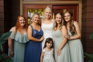 Bride in white gown poses with bridesmaids in sage green dresses and mother in blue at outdoor wedding.