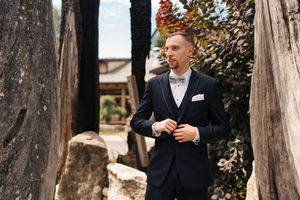 Elegant groom in navy suit and bow tie posing outdoors among rustic stone pillars.