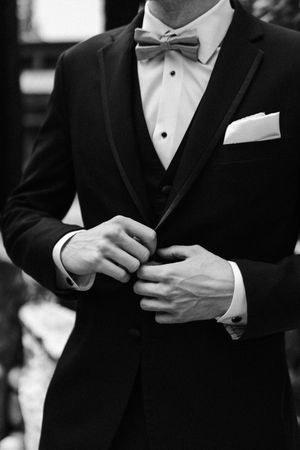 Black and white photo of a groom in a tuxedo with bow tie and pocket square, buttoning his jacket.