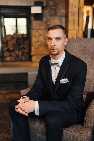 Elegant groom in black tuxedo with bow tie sits in armchair near rustic stone fireplace with stacked wood.
