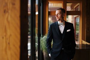 A dapper groom in a navy suit and bow tie stands by large windows, smiling confidently in an elegant venue.
