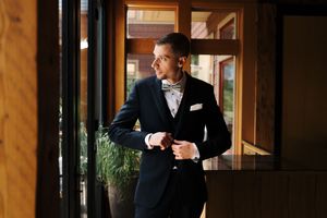 Elegant groom in navy suit and bow tie adjusting jacket cufflinks near window in upscale venue.