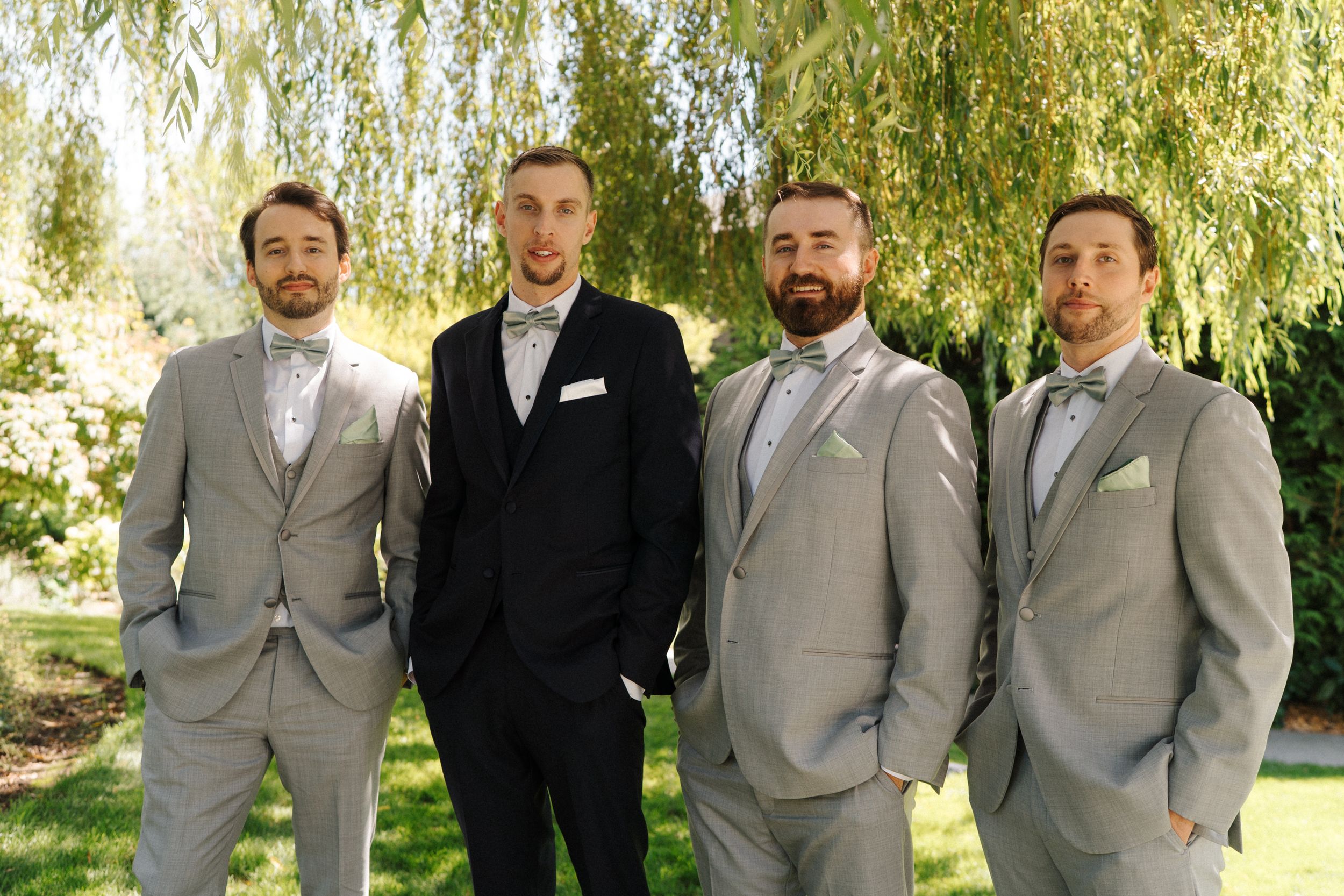 Groom in black suit poses with three groomsmen in grey suits and bow ties outdoors under willow tree.