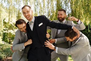 Groom in black suit laughing with three groomsmen in gray suits playfully surrounding him outdoors.