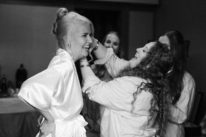 Bridesmaids in white robes laughing and helping bride with makeup during wedding preparations, black and white photo.