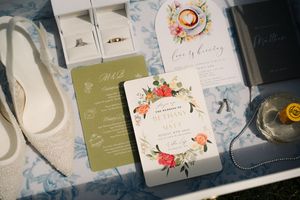 Elegant wedding flat lay featuring floral invitations, bridal heels, rings, and stationery on a marble surface.