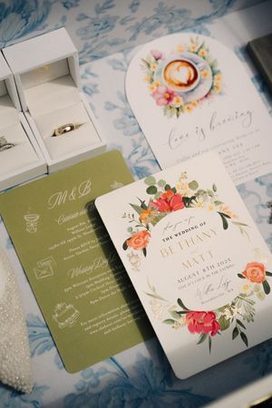 Elegant wedding invitation suite featuring floral designs, gold accents, olive green card, and a coffee-themed insert on blue fabric.