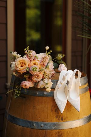 White lace bridal heels and a blush pink rose bouquet displayed on a rustic wooden wine barrel.