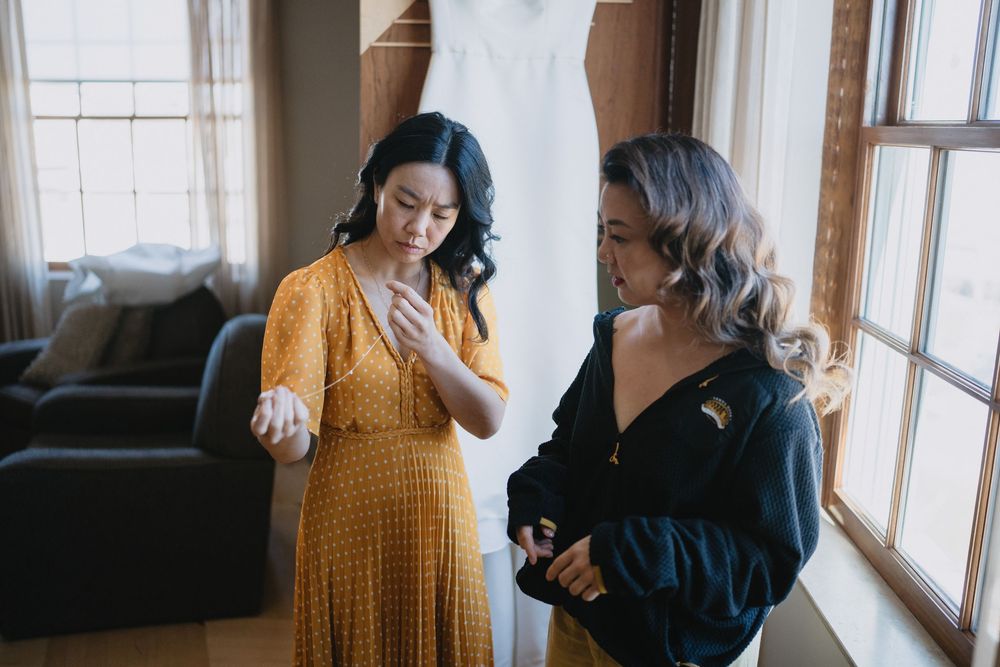 Two women in a sunlit room, one in yellow dress examining jewelry before a wedding ceremony.