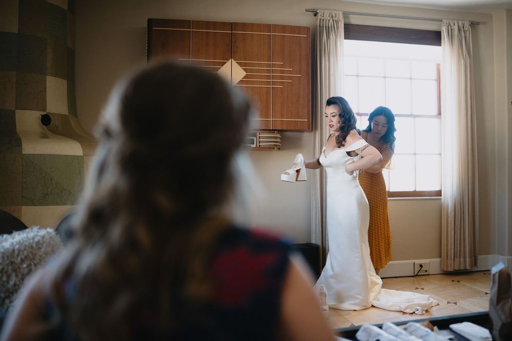 Bridesmaid helps bride with her wedding dress while another guest watches in a warmly lit room.