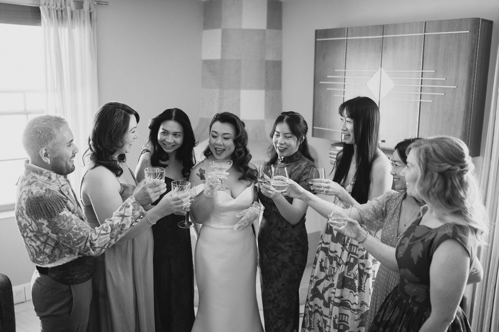 Black and white photo of bride in fitted gown toasting champagne with six bridesmaids in hotel room.