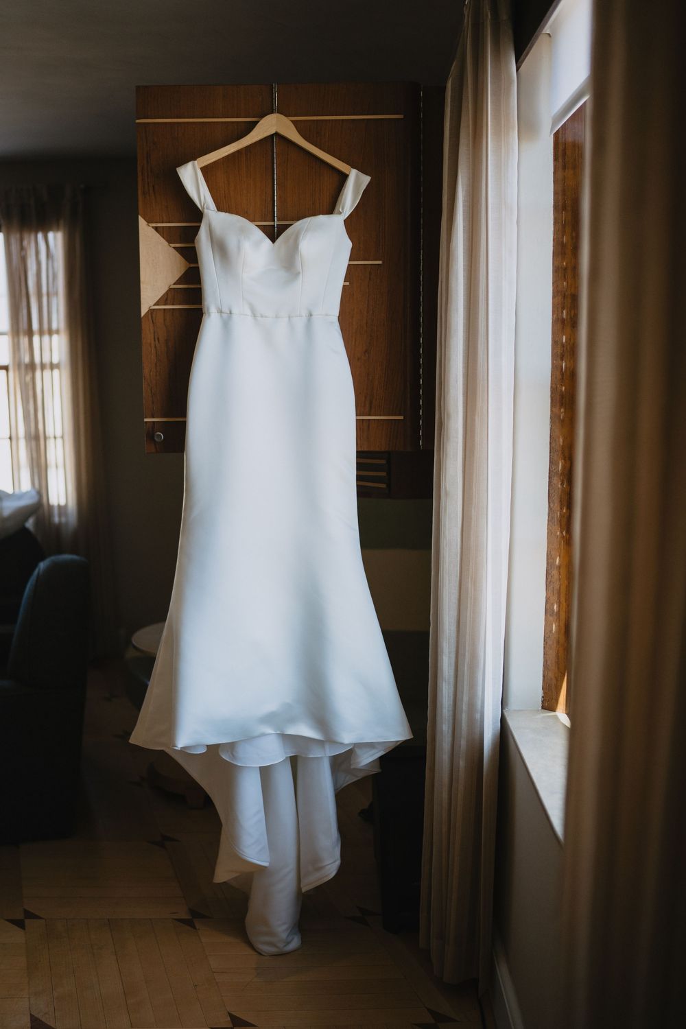 Elegant white mermaid wedding dress with sweetheart neckline hanging on wooden hanger by hotel window.