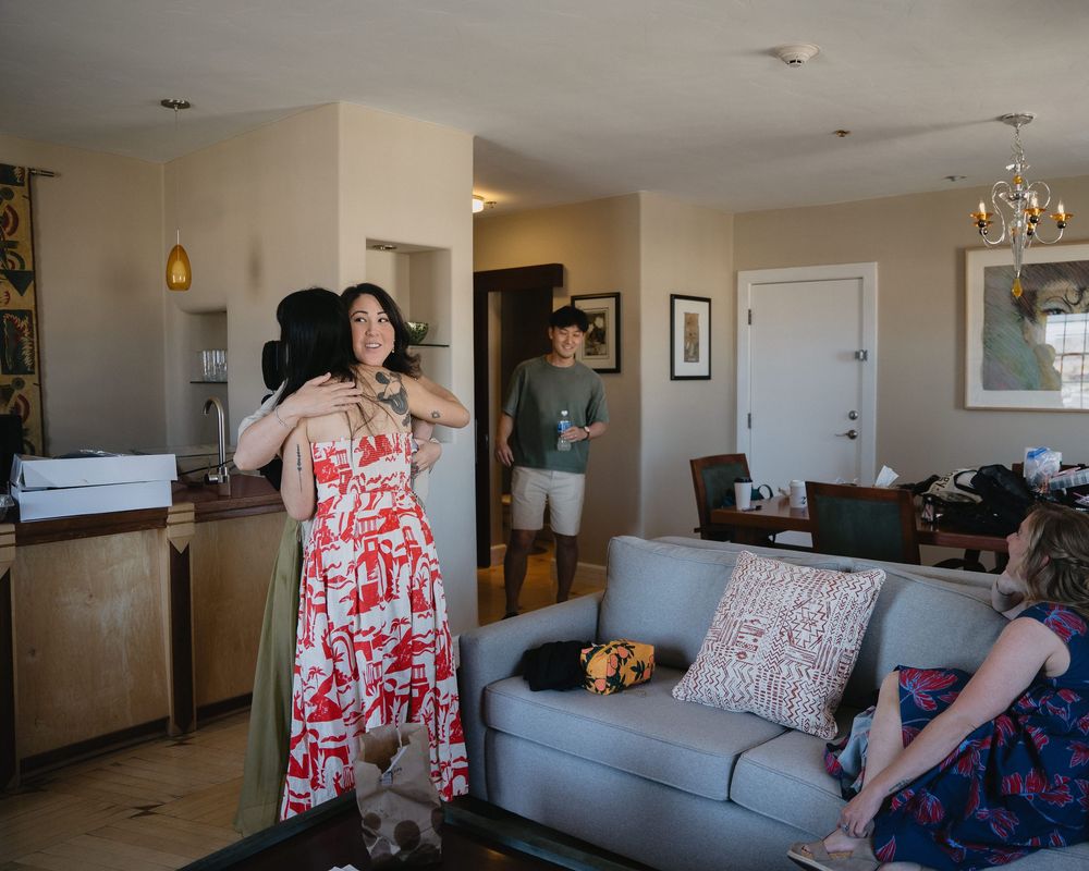 Woman in floral red dress showing outfit to friends in modern hotel suite before wedding celebration.