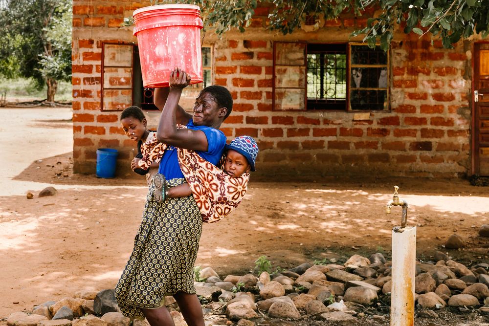 African woman carrying baby on back and red bucket on head near brick building in rural village.