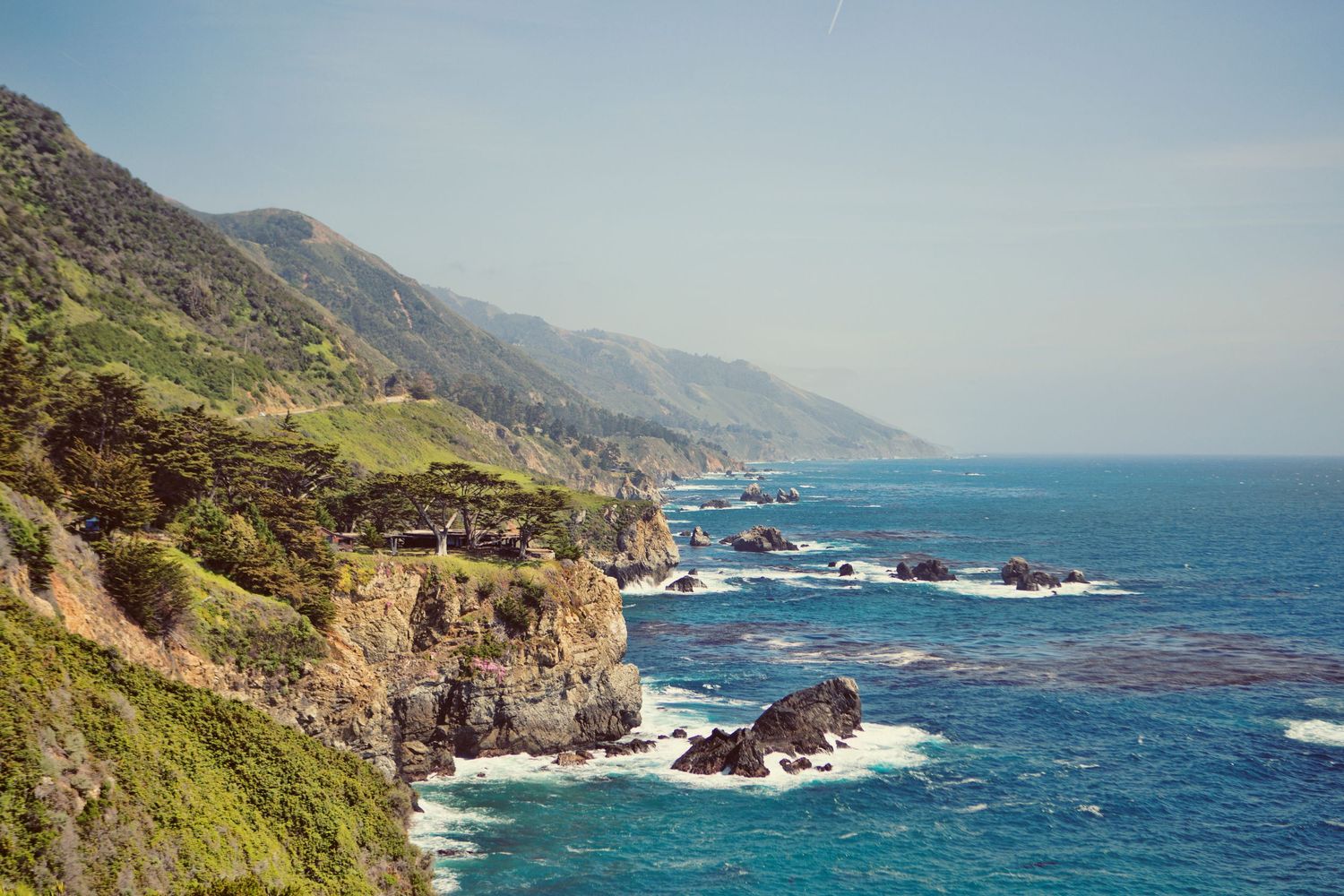Rocky coastal cove with turquoise waves, sandy beach, and lush green cliffs overlooking the Pacific Ocean.