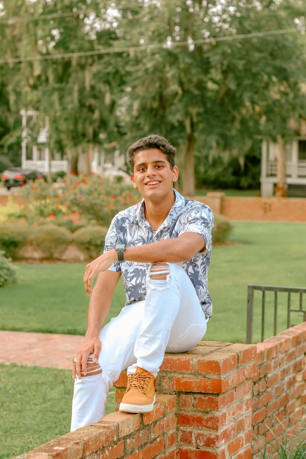 A casual portrait of a person in a floral shirt sitting on a brick wall in a garden setting.