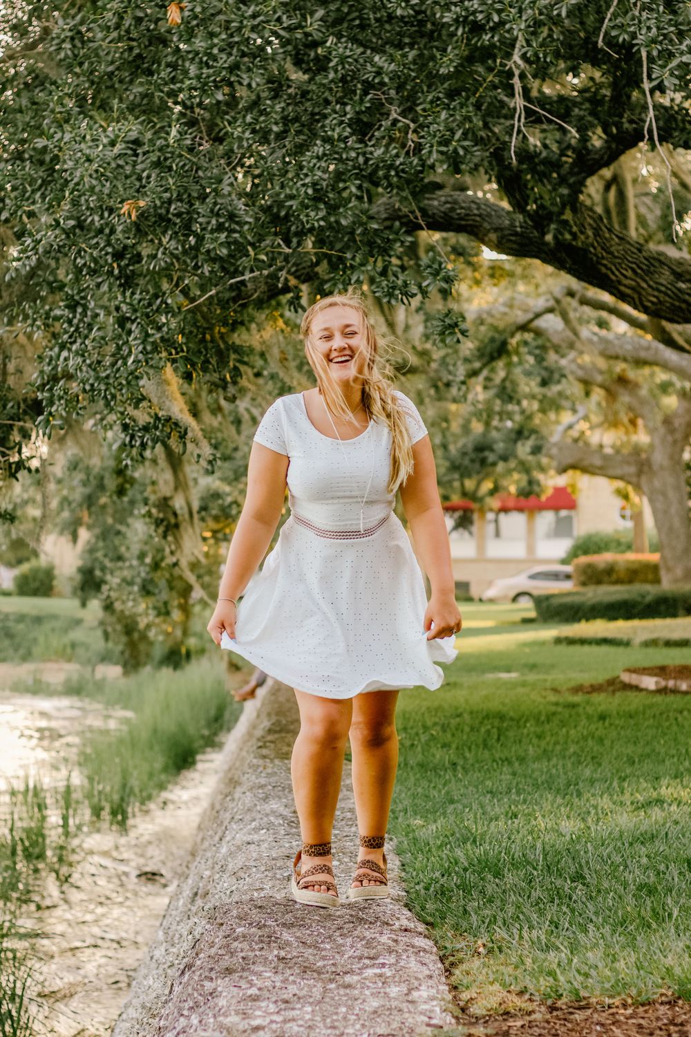Person in white dress twirling joyfully on path under tree canopy.