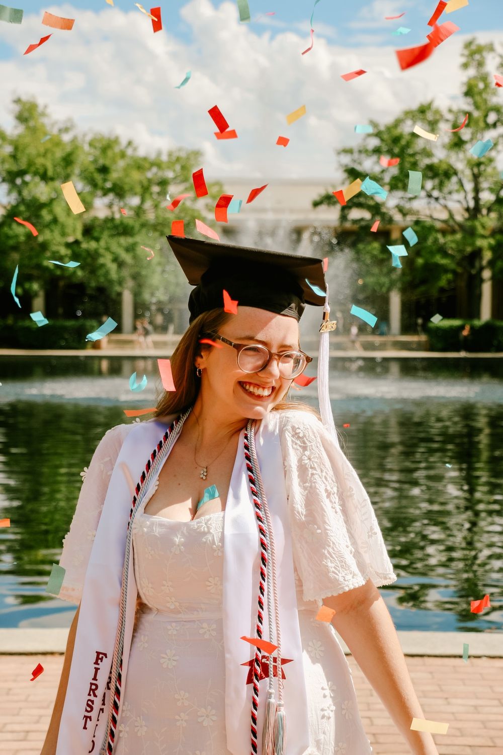 Graduate celebrating with confetti flying against blue sky and water fountain backdrop.