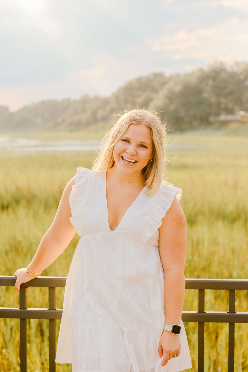A person in a white dress stands by a railing overlooking a marsh at sunset.