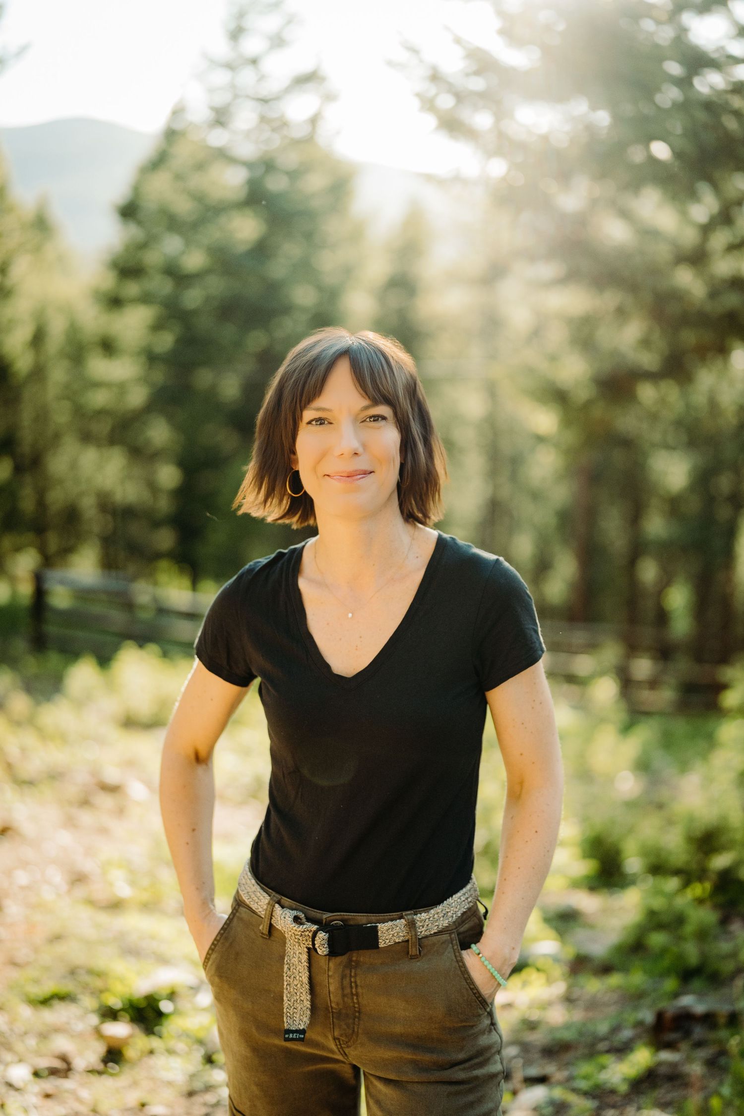 Woman in black t-shirt and olive pants smiling outdoors in a sunlit forest setting for a headshot photo by McGlone Photography