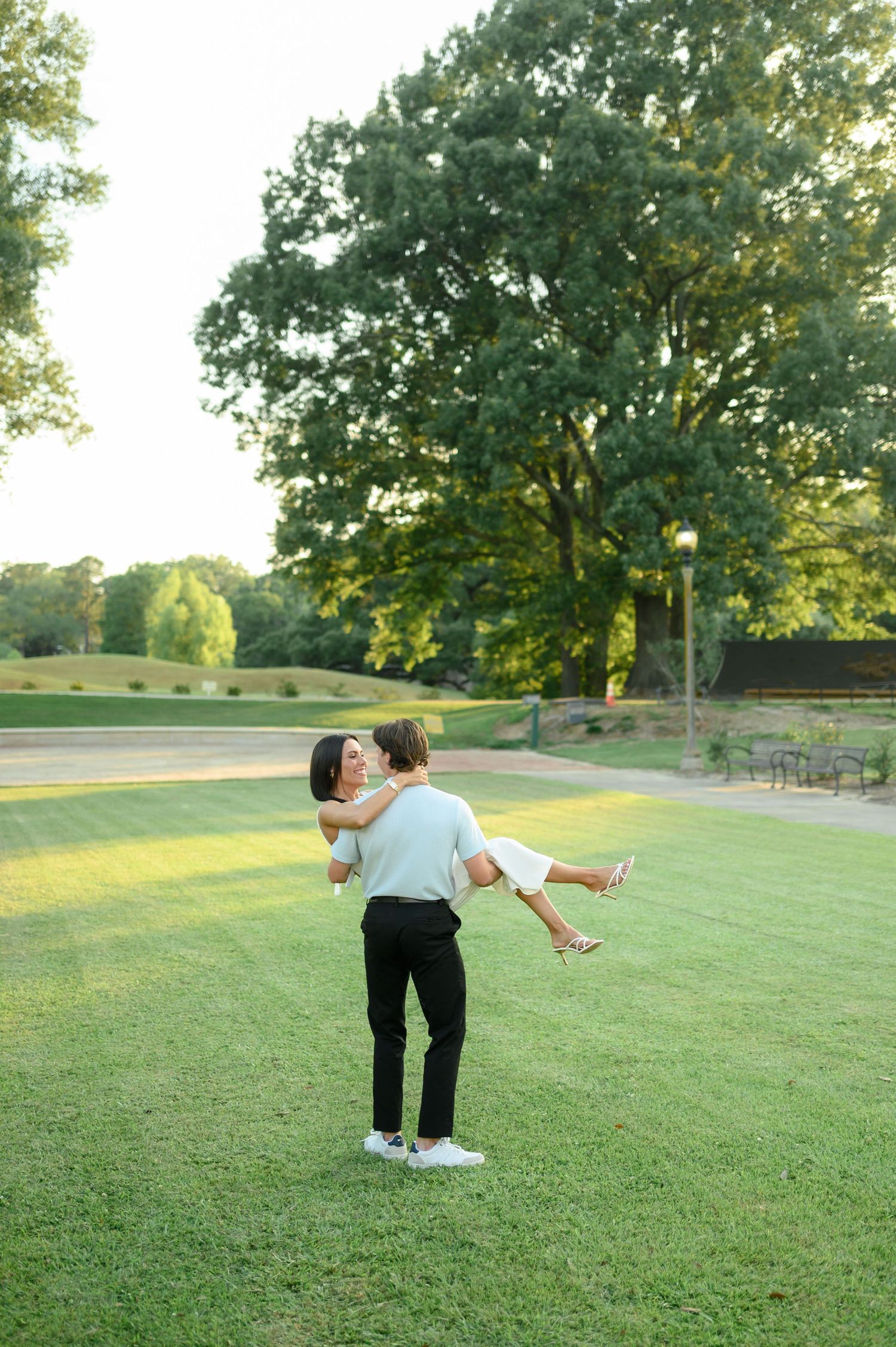 Man lifting woman in a romantic embrace on a lush green lawn with large trees in the background.