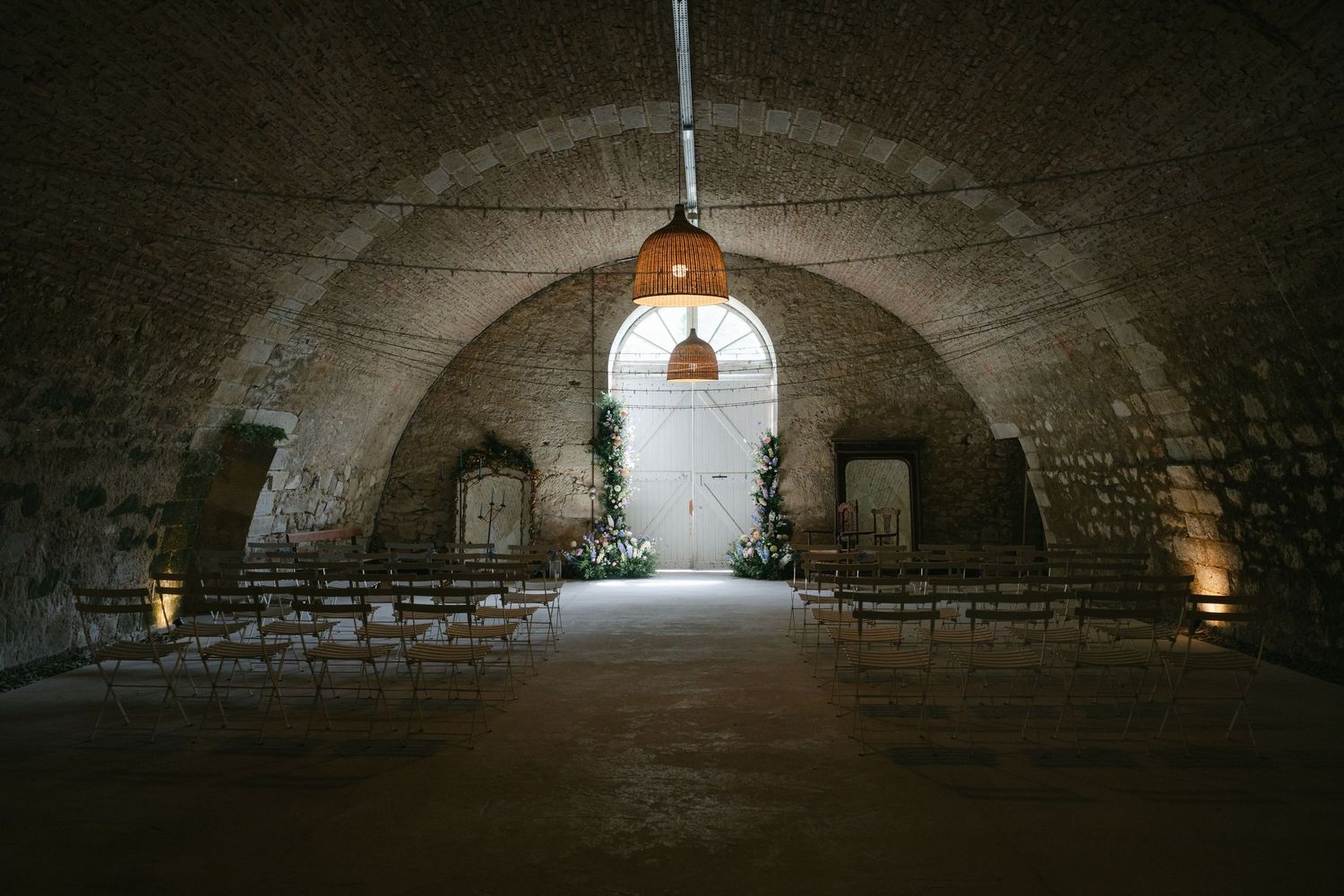 Stone vaulted ceiling wedding ceremony venue with arched architecture, wooden chairs, and warm pendant lighting.