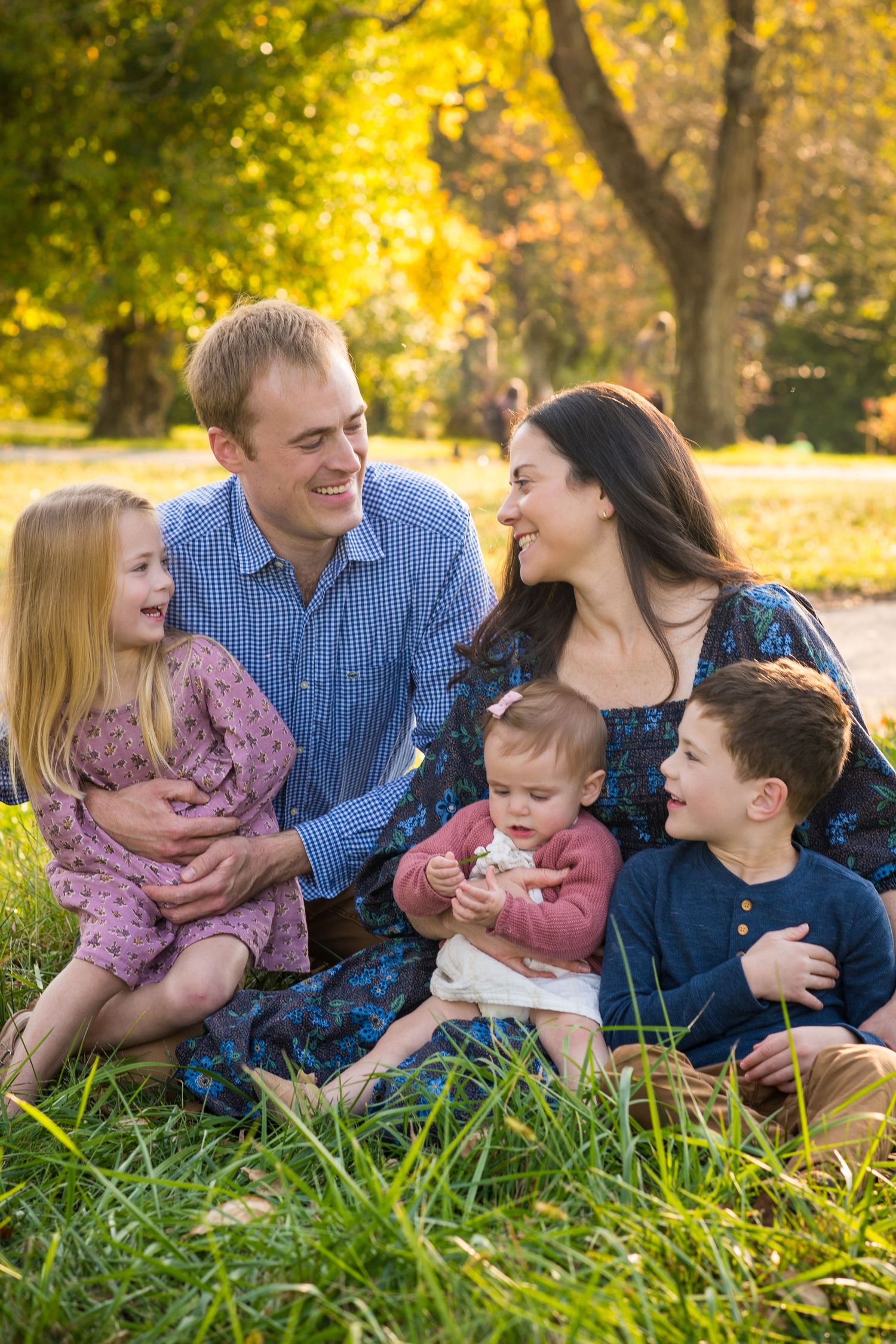 Happy family of five sitting on grass in autumn park, parents smiling at each other with three young children.