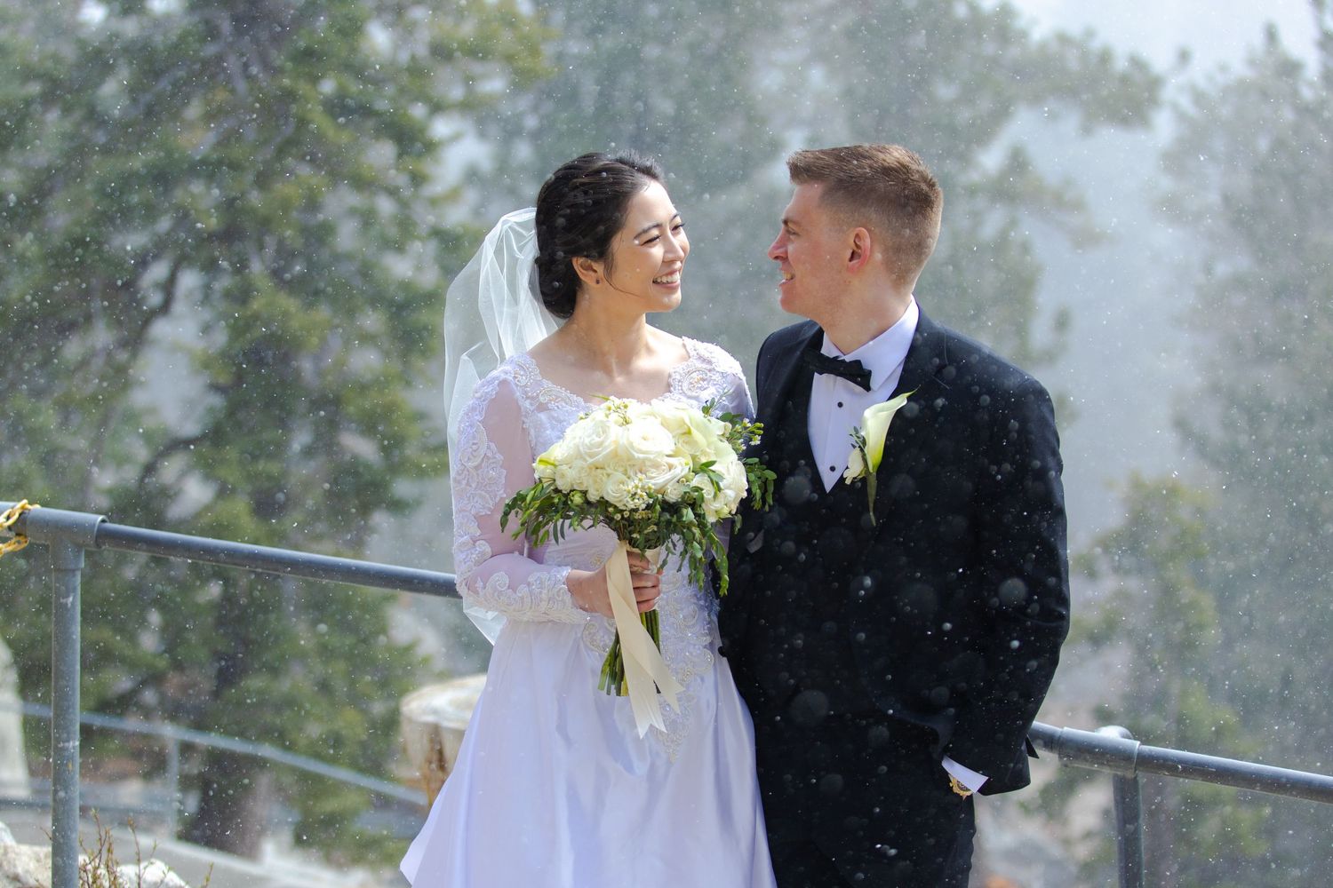 A bride and groom share a romantic moment outdoors in light snowfall, she holds white bouquet in wedding dress.