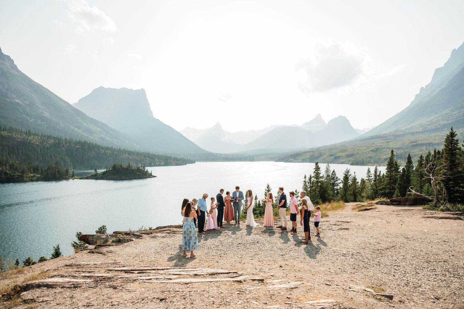 Wedding party gathered on rocky overlook above a pristine mountain lake surrounded by peaks in Glacier National Park.