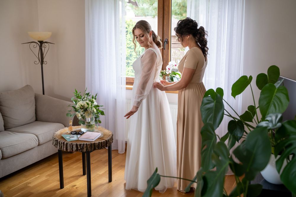 Dwie kobiety w eleganckich sukienkach stoją w jasnym pokoju udekorowanym roślinami doniczkowymi, przygotowując się na specjalną okazję.